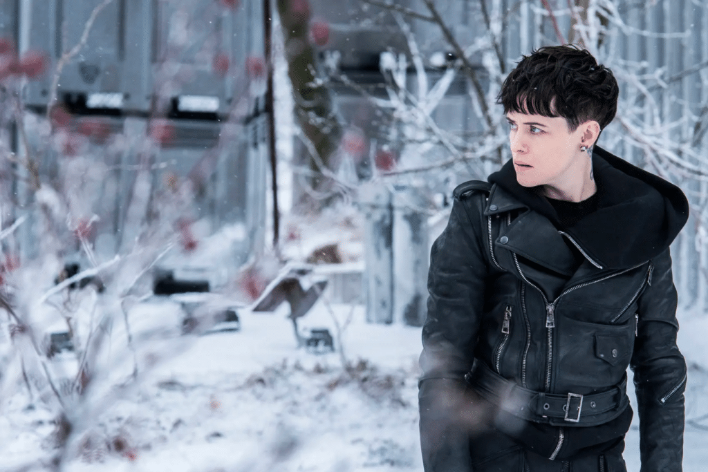 Image of Claire Foy in the role of Lisbeth Salander in 'Girl in the Spiders Web'. She is wearing a leather jacket, is surrounded by snow and is looking at something off-camera.